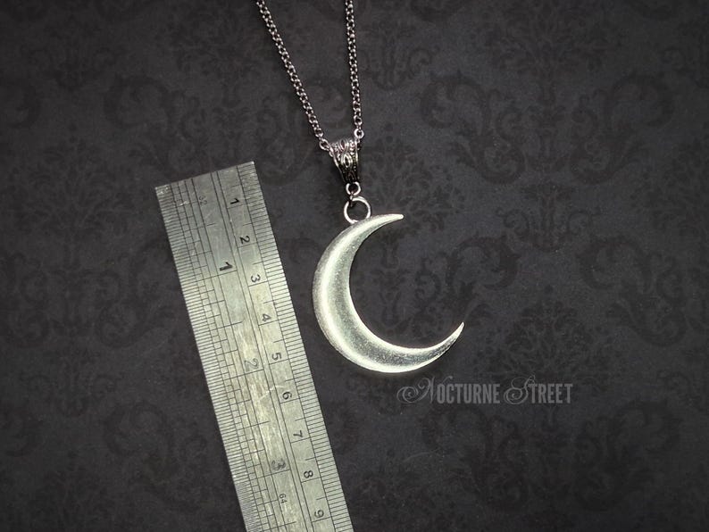 Silver crescent moon pendant lying on a dark gothic damask background next to a steel ruler. It shows the length of the moon and connector to measure approximately 6cm.