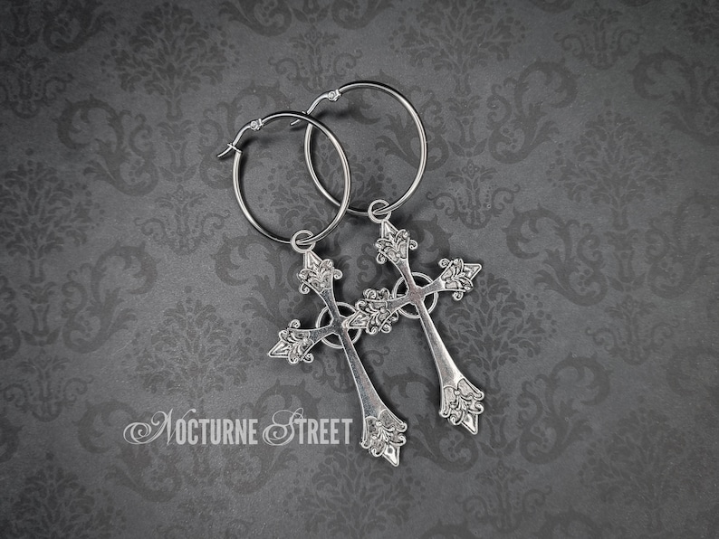A pair of Stainless Steel hoop earrings with large ornate crosses hanging from them, lying on a grey damask background.