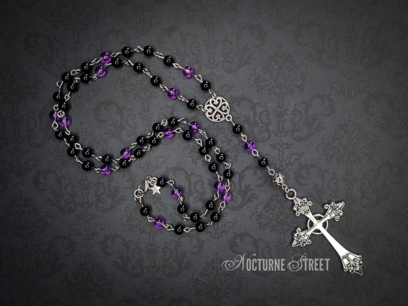 A gothic rosary lying on a grey damask background. The rosary is made of purple and black glass beads, has a celtic knot in the centre and has a decorative cross hanging from the end.