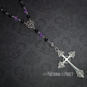 May include: A silver rosary necklace with black and purple beads and a silver cross pendant. The cross has a detailed design and is hanging from a silver chain.