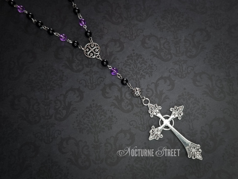 May include: A silver rosary necklace with black and purple beads and a silver cross pendant. The cross has a detailed design and is hanging from a silver chain.