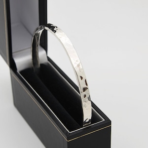 Könnte beinhalten: Ein silbernes, gehämmertes Armreif-Armband in einer schwarzen Samtbox. Das Armband ist ein schlichtes, elegantes Design.