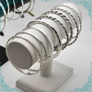 May include: A set of silver bangles, some with a hammered finish and others with a twisted rope design, displayed on a white stand.
