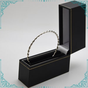 May include: A silver twisted bangle bracelet in a black velvet lined jewelry box with a gold trim.