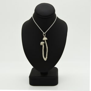 Könnte beinhalten: Eine silberfarbene Anhängerkette mit einem stilisierten Pilzdesign. Der Anhänger zeigt zwei Pilzfiguren, eine größere und eine kleinere, die an einer filigranen Kette hängen. Die Halskette wird auf einer schwarzen Schmuckbüste vor weißem Hintergrund präsentiert.