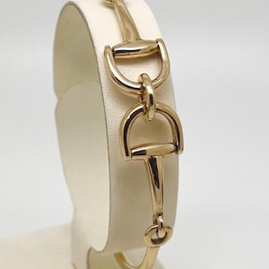 May include: A gold-toned bracelet featuring a horse bit design. The bracelet is displayed on a white stand, showcasing its polished finish and intricate details. The equestrian-inspired jewelry piece has a classic and elegant aesthetic.
