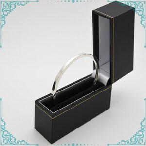 May include: A silver bangle bracelet in a black velvet lined box with a gold trim.