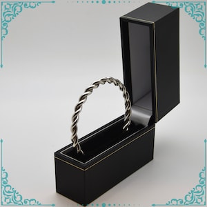 May include: A silver twisted bangle bracelet in a black velvet jewelry box with a gold trim.