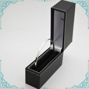 May include: A silver bangle bracelet in a black velvet lined box. The box is open and the bracelet is sitting inside.