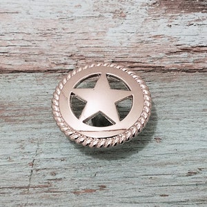 May include: A silver-colored metal button with a star design. The star is in the center of the button, surrounded by a circular border with a rope-like texture. The button is likely for clothing or accessories.