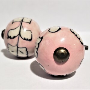 May include: Two pink ceramic knobs with white and black floral designs. The knobs have a brass screw on the back.