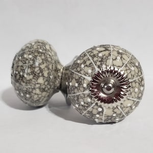 May include: Two ceramic door knobs with a white and gray speckled pattern. The knobs have a silver metal base with a decorative design.