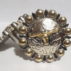 May include: Silver and gold cowboy buckle with a bull's head design. The buckle has a decorative border with gold accents.
