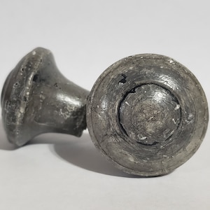 May include: A silver-toned metal knob with a round, slightly raised center and a smaller, round base. The knob has a worn, aged appearance.