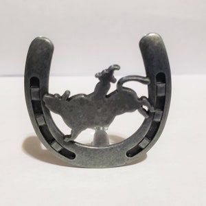 May include: A silver metal horseshoe with a cutout of a cowboy riding a bucking bull. The horseshoe is on a white background.