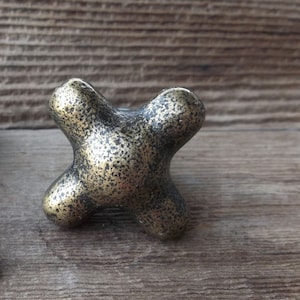 May include: A gold-toned metal cabinet knob with a unique, geometric design. The knob has a textured surface and a slightly distressed finish.