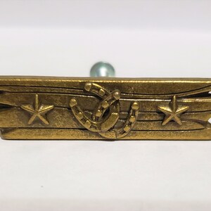 May include: Gold-toned metal drawer pull with a rustic design. It features two horseshoes and two stars, with a woodgrain texture.