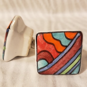 May include: Two decorative ceramic cabinet knobs. One is square with a colorful abstract design in red, orange, blue, and green. The other knob is white with a partial view of a similar design. Hardware is visible.