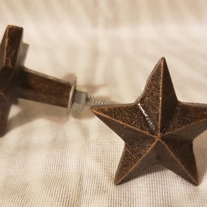 May include: Two bronze-colored star-shaped cabinet knobs. One knob is in focus, displaying a five-pointed star design. The other knob is slightly out of focus, showing the star shape and the screw hardware. The knobs have a rustic, vintage aesthetic.