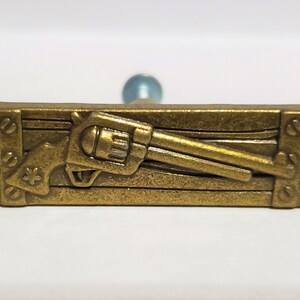May include: A gold-toned metal pin with a detailed design of a revolver pistol on a wooden plank. The pin has a textured surface and a rustic look.