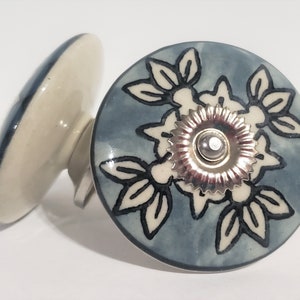 May include: Two ceramic cabinet knobs. One knob is in the foreground, displaying a floral design with a silver center on a blue background. The other knob is in the background, showing a white and blue design. These knobs are suitable for furniture.