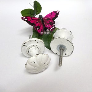 May include: Two white ceramic cabinet knobs with a distressed finish. The knobs have a scalloped design and are attached to a metal screw. A pink butterfly with black markings is in the background.