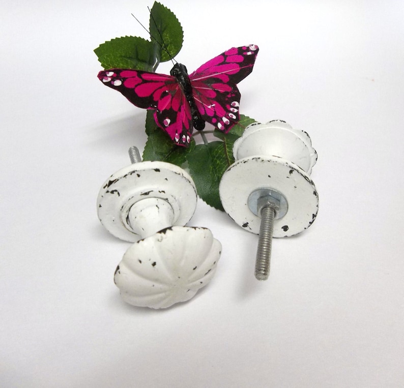 Może przedstawiać: Dwa białe ceramiczne gałki do szafek z wykończeniem w stylu vintage. Gałki mają falisty wz&oacute;r i są przymocowane do metalowej śruby. Na tle znajduje się r&oacute;żowy motyl z czarnymi oznaczeniami.