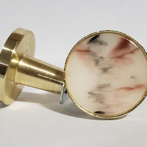 May include: A decorative brass seal stamp with a round, marbled design in cream, black, and red. The stamp has a gold-colored metal rim and a brass handle. The seal is designed for adding a unique touch to crafts and stationery.