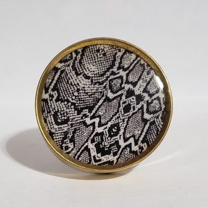 May include: A round cabinet knob with a snakeskin pattern in black and white, set within a gold-colored metal frame. The knob is a decorative hardware element, likely for furniture or cabinetry.