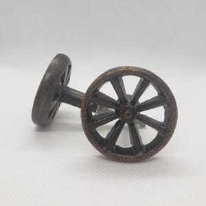 May include: Two bronze-colored cabinet knobs in the shape of wagon wheels. Each wheel has a central hub with spokes radiating outwards. The knobs are mounted on a metal stem, suitable for furniture or decorative use.