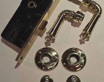 May include: A collection of antique brass door hardware, including a rectangular lock with a black face, two curved door handles, two round doorknob backplates, and two oval keyhole covers. The hardware is arranged on a light-colored surface.