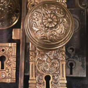 May include: Close-up of antique brass door hardware, including a detailed doorknob with floral patterns and a matching escutcheon. The hardware has an aged, vintage appearance, with intricate carvings and keyholes. Other hardware pieces are visible.