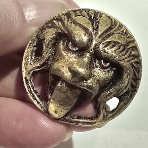 May include: A circular metallic button with a gold-toned finish featuring a relief sculpture of a lion head with a protruding tongue. The design includes textured mane details and two small circular holes for attachment on the right and left sides.