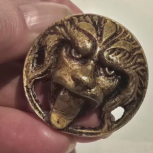 May include: A round, antique-style button with a detailed relief of a lion's face. The button is made of a gold-colored metal and features intricate detailing of the lion's mane and facial features. The button has a hole for sewing.