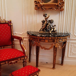 Può includere: Scena d'interni di casa delle bambole con una poltrona e un pouf rossi, entrambi con dettagli dorati e tessuto fantasia. Un tavolino con piano in marmo e un ritratto incorniciato completano l'arredamento classico.