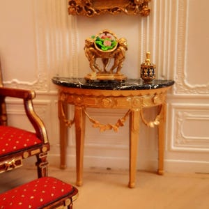 May include: Miniature gold-painted furniture set, including a table with a black marble top, a chair, and a footstool, all with red upholstery. A framed portrait and a decorative gold object are on the table.