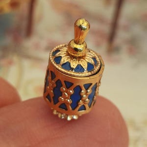 May include: A miniature gold-colored container with a blue enamel floral design. The container has a lid and a small handle. The container is held between two fingers.