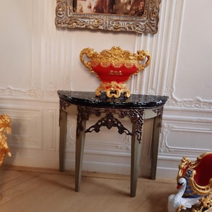 Könnte beinhalten: Eine Miniatur-Zierurne in Rot und Gold mit aufwendigen Details steht auf einem kleinen Tisch mit schwarzer Platte und dekorativen Beinen. Der Tisch steht vor einer weißen Wand mit einem gerahmten Bild.