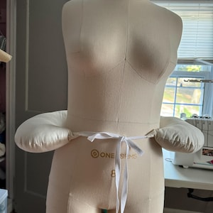 Könnte beinhalten: Eine weiße Schneiderpuppe mit zwei weißen Stoffpolstern, die an der Taille befestigt sind. Die Polster sind mit einem weißen Band zusammengebunden. Die Schneiderpuppe hat den Text "ONE DMS" auf der Vorderseite.