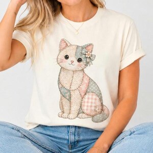Puede incluir: Una camiseta de manga corta de color crema con un gráfico central de un gato sentado. El gato está diseñado con una estética de patchwork, incorporando varios patrones suaves como lunares y cuadros vichy en tonos rosa, azul y beige.