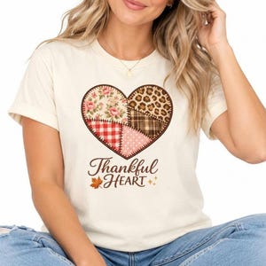Könnte beinhalten: Ein cremefarbenes Kurzarm-T-Shirt mit einem großen herzförmigen Aufdruck, der aus verschiedenen Patchwork-Mustern wie Blumen, Leopardenmuster, Karos und Punkten besteht. Unter dem Herzen steht der Schriftzug Thankful Heart in Braun.