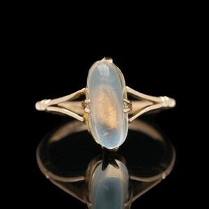 May include: A gold ring with an oval, translucent, light blue gemstone. The ring's band has a delicate, openwork design. The gemstone has a subtle, golden hue in the center, set against a black background.