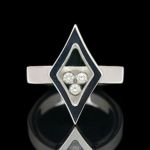 May include: A silver ring featuring a diamond-shaped design with three round diamonds. The ring has a sleek, modern aesthetic, with a polished finish and a dark background.