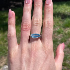 K&ouml;nnte beinhalten: Ein silberner Ring mit einem rechteckigen, hellblauen Edelstein. Der Ring befindet sich an einer Hand mit rosa Nagellack. Der Hintergrund ist verschwommen, mit gr&uuml;nem Gras und Sonnenlicht.