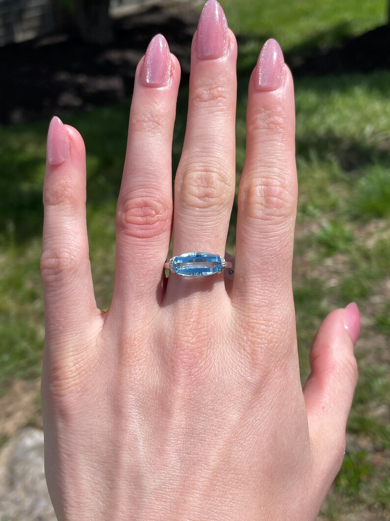 K&ouml;nnte beinhalten: Ein silberner Ring mit einem rechteckigen, hellblauen Edelstein. Der Ring befindet sich an einer Hand mit rosa Nagellack. Der Hintergrund ist verschwommen, mit gr&uuml;nem Gras und Sonnenlicht.