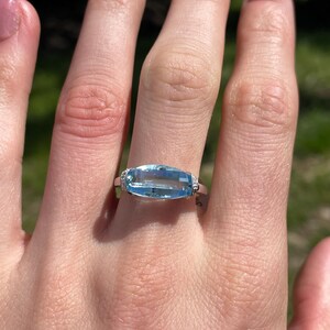 K&ouml;nnte beinhalten: Ein rechteckiger, hellblauer Edelsteinring mit kleinen, klaren Akzentsteinen auf einem silbernen Band. Der Ring befindet sich an einer Hand mit einem verschwommenen gr&uuml;nen Hintergrund.