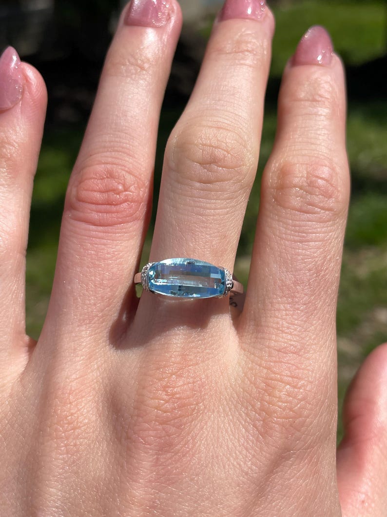 K&ouml;nnte beinhalten: Ein rechteckiger, hellblauer Edelsteinring mit kleinen, klaren Akzentsteinen auf einem silbernen Band. Der Ring befindet sich an einer Hand mit einem verschwommenen gr&uuml;nen Hintergrund.