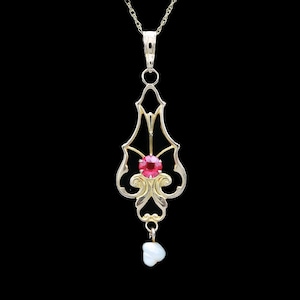 May include: A gold-toned pendant necklace with an ornate, openwork design. The pendant features a central, round, red gemstone and a small, white, teardrop-shaped pearl. The necklace hangs against a black background.
