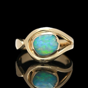 May include: A gold ring featuring a central, oval-shaped opal gemstone with vibrant blue and green iridescent flashes. The polished gold band has a smooth, curved design that frames the stone, creating an eye-like shape. The ring is set against a black background.