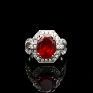 May include: A silver ring with a large, oval-shaped ruby surrounded by a halo of smaller diamonds. The ring has a delicate, intricate design.